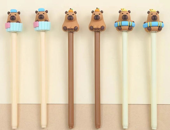 A set of cute gel pens designed by BC Mini featuring capybara toppers. The pens come in various colors with adorable capybaras sitting on decorative elements like cupcakes and lifebuoys.