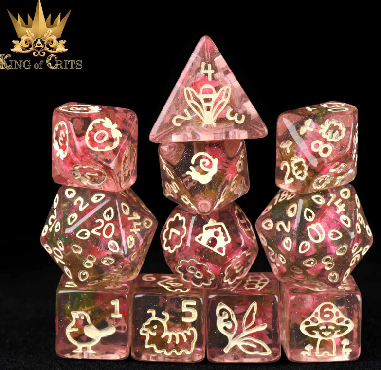 A colorful 11-piece RPG dice set from King of Crits featuring cozy cottagecore designs. The dice are made of translucent resin and showcase intricate illustrations of nature, including flowers, animals, and whimsical symbols. The set includes various shapes like a pyramid and D20 dice, all intricately detailed and shimmering with hints of glitter.