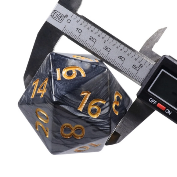 The 55mm Titan D20 - Black Pearl by Foam Brain Games is a striking twenty-sided die featuring a marbled black and gray finish with vibrant gold numbers. It is being measured with a digital caliper, showcasing its size and detail.
