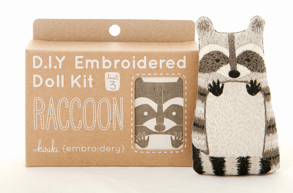 Raccoon Doll Embroidery Kit from Kiriki Press featuring a charming raccoon-shaped plush doll and a cardboard box with DIY embroidery instructions. The kit is labeled as level 3, indicating a moderate difficulty for crafters to create their own embroidered raccoon.