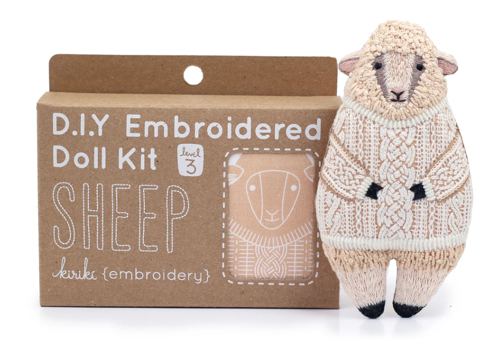 A DIY Sheep Doll Embroidery Kit by Kiriki Press, featuring a detailed embroidered sheep doll in a cozy sweater. The packaging highlights that it's a level 3 kit for skilled crafters. The doll is crafted from plush textile with intricate stitching, perfect for crafting enthusiasts.