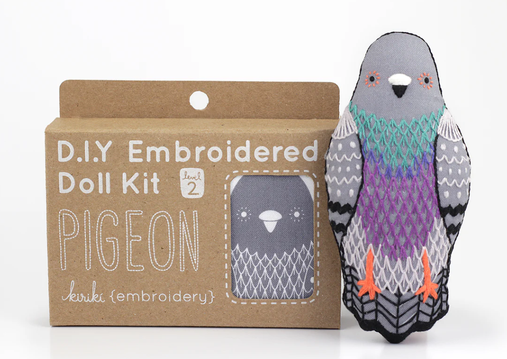 A Pigeon Doll Embroidery Kit by Kiriki Press, featuring a colorful embroidered pigeon plush. The kit is labeled as a DIY project, level 2. The packaging is a brown cardboard box displaying the pigeon design.