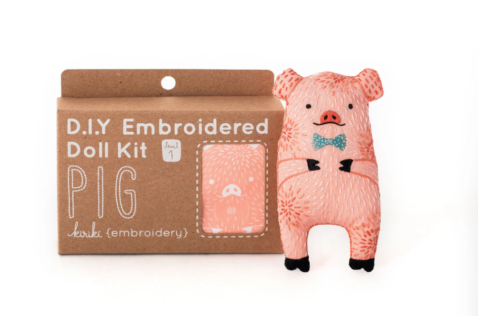 The Pig Doll Embroidery Kit by Kiriki Press features a charming pink pig plush doll adorned with a blue bowtie, showcasing its embroidered details. The kit is packaged in a light brown box labeled 'D.I.Y Embroidered Doll Kit' with a level 1 difficulty rating, perfect for beginner crafters looking to create their own adorable pig character.