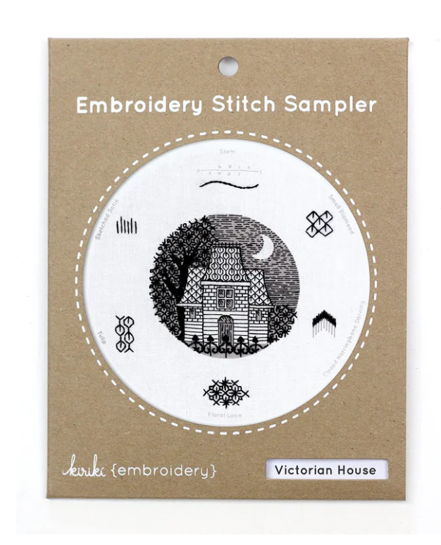 A Victorian House embroidery stitch sampler kit by Kiriki Press. The design features a detailed illustration of a Victorian house surrounded by trees and decorative stitching patterns. Packaged in a stylish cardboard cover with the words 'Embroidery Stitch Sampler' at the top, this DIY kit provides everything needed for embroidery enthusiasts.