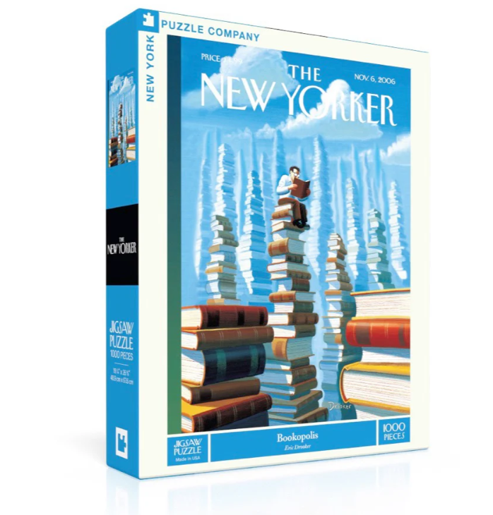 BOOKOPOLIS jigsaw puzzle by New York Puzzle Company. Features a whimsical illustration of a man reading atop towering stacks of books against a vibrant blue background. The packaging includes the title, artist Eric Drober, and details of the puzzle with 1000 pieces.