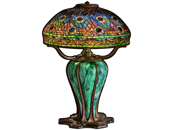 The Peacock Library Lamp features an elegant design with a vibrant stained glass shade adorned with peacock feathers in shades of blue, green, and orange. The lamp's base is crafted in a rich green color with a decorative, vintage-style metal finish, adding a touch of sophistication to any space. Ideal for enhancing mood and ambiance while serving as a stylish decorative piece.
