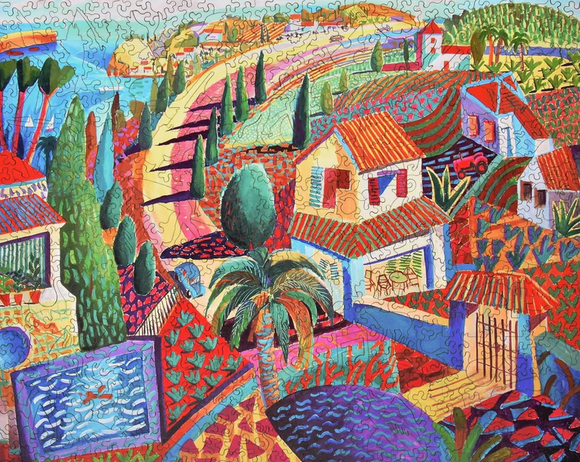 Vibrant jigsaw puzzle titled 'Spanish Coast' by Liberty Puzzles. The artwork features a colorful depiction of a coastal landscape with rolling hills, palm trees, charming houses with red roofs, and a winding pathway leading toward the sea. The scene is filled with rich colors and intricate details, perfect for puzzle enthusiasts.