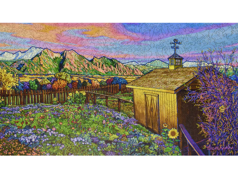 A vibrant jigsaw puzzle titled 'Changing Light, Changing Seasons' featuring a scenic landscape. The image includes colorful flowers in a lush garden, a quaint wooden shed, and majestic mountains in the background, all framed by a pink and orange sunset sky, creating a serene and inviting atmosphere.