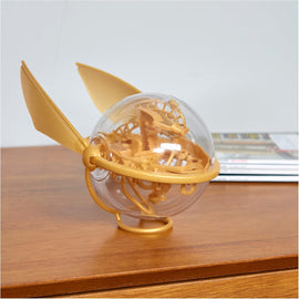 Harry Potter Perplexus Golden Snitch mechanical puzzle by Spin Master. The puzzle features a transparent orb with intricate internal mechanisms and golden wings, placed on a wooden surface.
