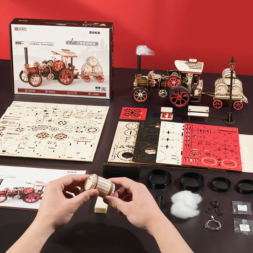 A Steam Engine DIY kit by Robotime displayed on a table. The kit includes intricately designed wooden pieces, tools, and components for assembly. A person holds a wooden barrel part with the assembled steam engine in the background, showcasing its detailed gears and wheels, underlining the creativity and craftsmanship involved in building this mechanical model.