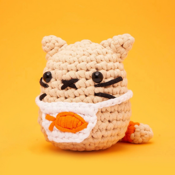 A cute crocheted cat named Nico, made from soft yarn. Nico has a round body with distinct facial features, including large black eyes, whiskers, and tiny ears. It is adorned with a white apron featuring an orange fish design, set against a bright orange background.