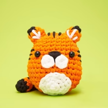 A cute crocheted toy featuring an orange tiger with black stripes, white facial features, and round black eyes, set against a bright green background. This DIY crochet kit offers a beginner-friendly project for crafting enthusiasts.