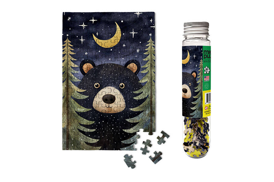 A whimsical jigsaw puzzle titled 'Forest Bear Wildlife Micropuzz.' The image features a cute black bear against a nighttime forest backdrop with trees and a crescent moon. The puzzle pieces are presented in a clear tube, showcasing the colorful shapes. A few scattered pieces are shown alongside the puzzle, inviting engagement.