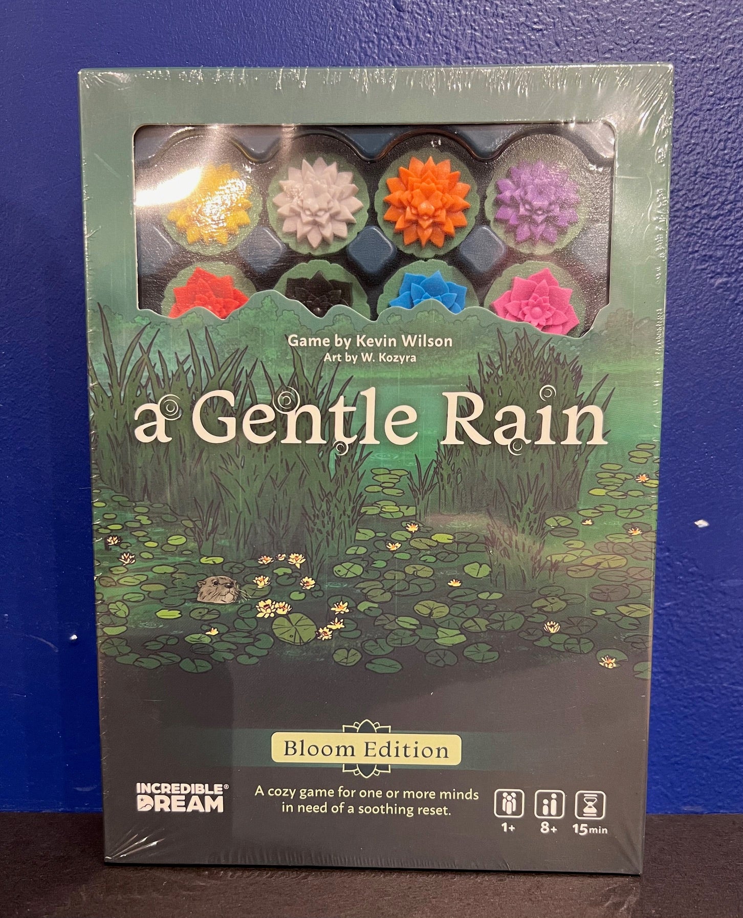 The game 'A Gentle Rain' by INcredible Dream, featuring a captivating cover illustration of a serene pond with lily pads and colorful floral tokens. The box is brand new and displays the title prominently, along with game details indicating it is a cozy game for one or more players, ages 8 and up, with a playtime of approximately 15 minutes.