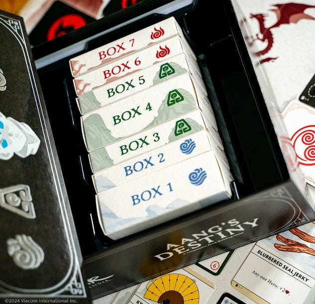 The Avatar Deck Builder: Aang's Destiny game, featuring a game box with seven smaller boxes labeled Box 1 through Box 7, each adorned with elemental symbols representing air, earth, fire, and water. The game components are spread on a table, highlighting the vibrant design and thematic elements related to the Avatar universe.