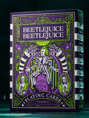 Beetlejuice Playing Cards designed by Theory 11, showcasing vibrant green and purple artwork featuring the iconic character. The packaging has intricate designs with skeletons and stripes, highlighting a blend of whimsical and spooky themes. Perfect for collectors and fans of the Beetlejuice franchise.