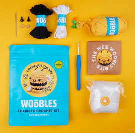 Cornelius the Bee Beginner Crochet Kit by Woobles, featuring colorful yarns in black, white, and yellow, safety eyes, crochet hook, stuffing material, and instructional booklet, all arranged on a bright yellow background.