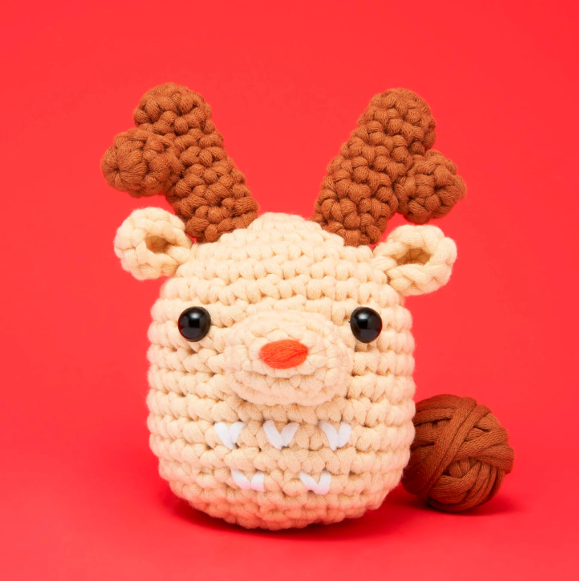 Ralph the Reindeer Beginner DIY kit by Woobles featuring a cute, plush reindeer with soft antlers and a warm smile. A ball of yarn is beside it, set against a vibrant red background.