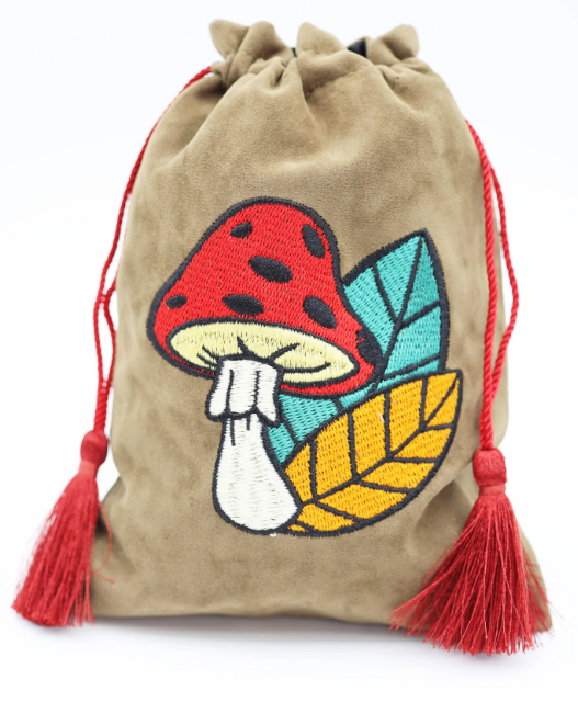A soft, suede dice bag featuring an embroidered design of a red mushroom with black spots and colorful leaves. The bag is cinched at the top with red tassels attached, perfect for storing gaming dice.