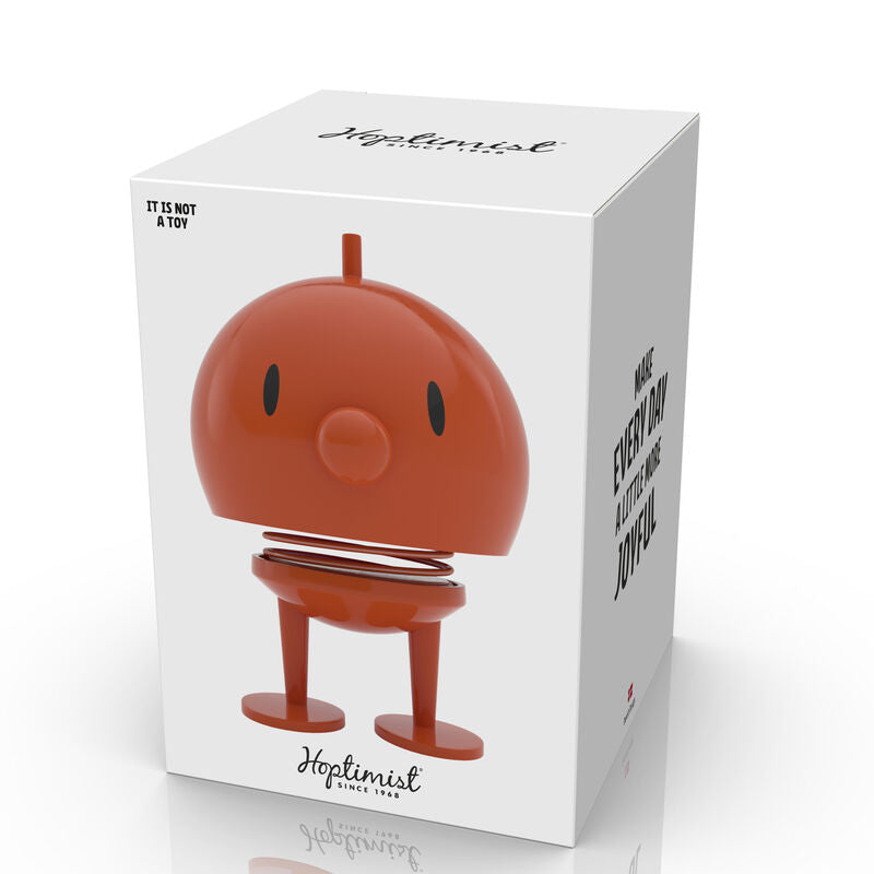 Hoptimist Bumble M in Orange, featuring a smiling face, spring body, and round head. The product is in a box with a minimalist design, emphasizing joyfulness.