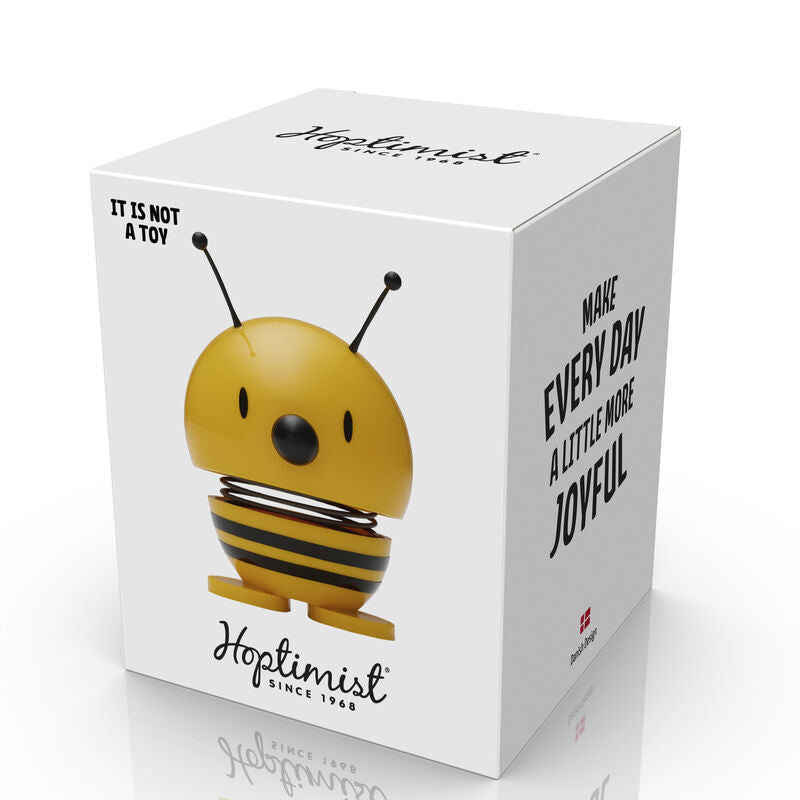 The Hoptimist Bee Yellow is a cheerful, spring-loaded figure designed to bring joy. It features a bright yellow body with black stripes, a smiling face, and antennae, all packaged in a white box displaying its playful message 'Make Every Day a Little More Joyful.' The packaging emphasizes that it is not a toy but rather a decorative item.