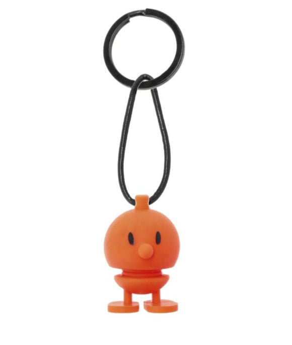 Hoptimist Keychain Bumble Oran features a cheerful orange bumble character with a round body and a smiling face, attached to a sturdy keyring via a black strap. The playful design is perfect for adding a touch of joy to your keys or bag.