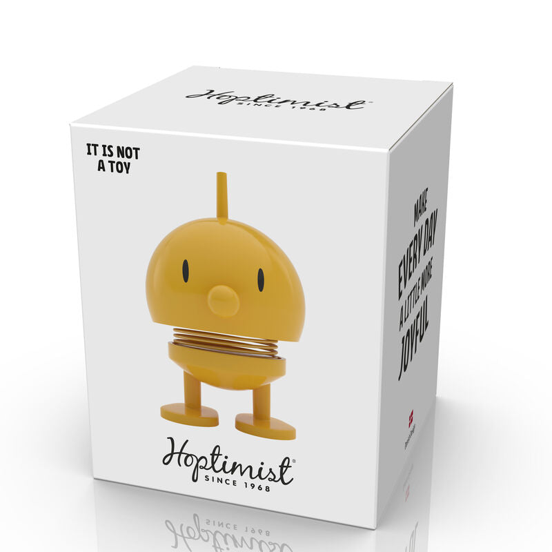 A Hoptimist Bumble L Yellow figure packaged in a box. The figure is a cheerful yellow character with a round head and small feet, featuring a smiling face. The box has a minimalistic design with the Hoptimist logo and text stating "It is not a toy." Perfect for adding a joyful touch to any space.