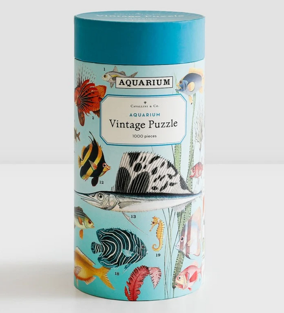 A cylindrical puzzle box for the Aquarium Vintage Puzzle by Cavallini & Co., featuring a light blue background decorated with vintage-style illustrations of tropical fish and sea life, topped with a solid teal lid.