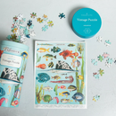 Vintage fish-themed jigsaw puzzle set with a printed puzzle sheet, a turquoise round tin, and scattered puzzle pieces on a light gray surface.