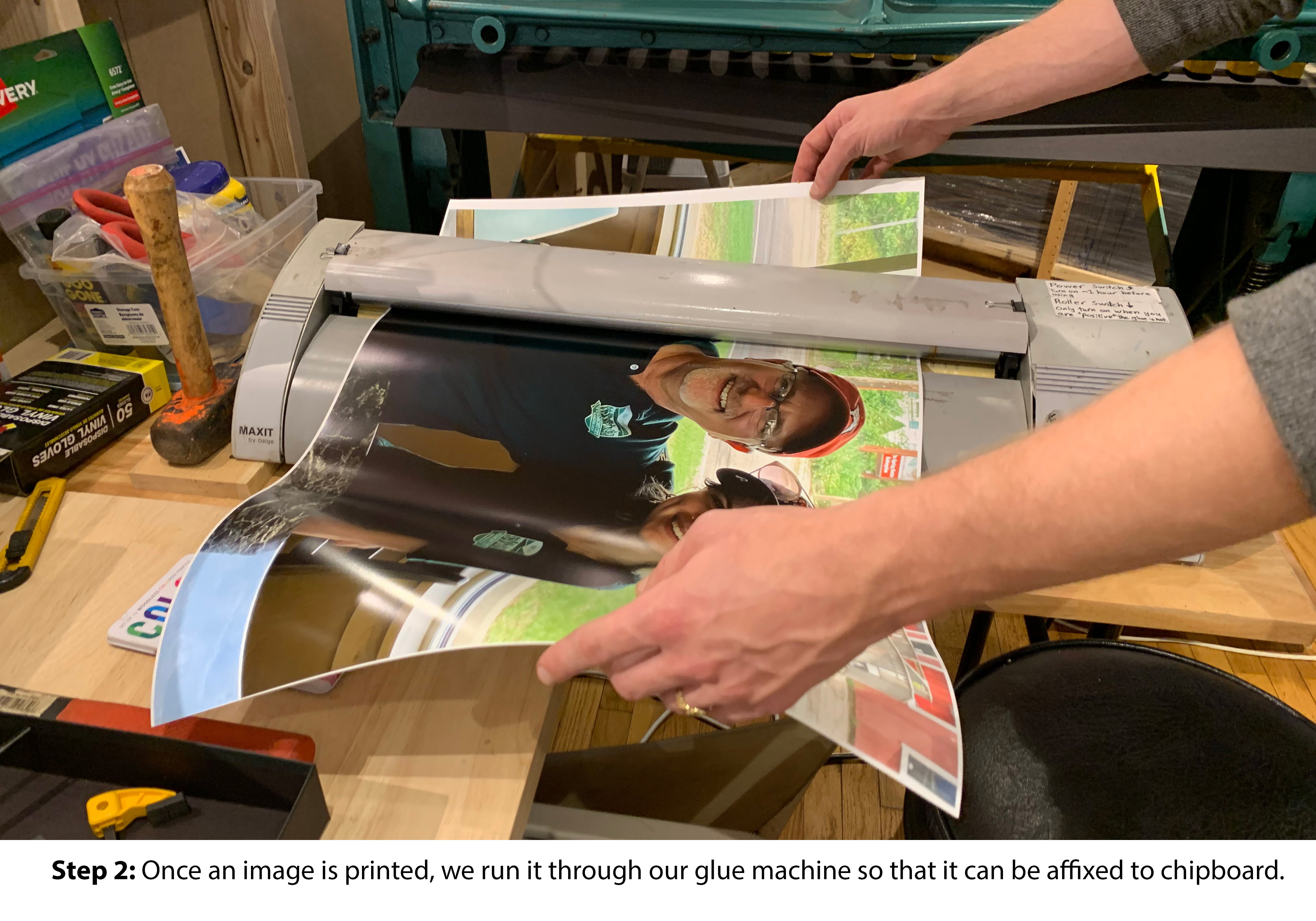 A person is operating a glue machine in a workshop, preparing to affix a printed image to chipboard. The setup includes tools and materials scattered on a work surface.