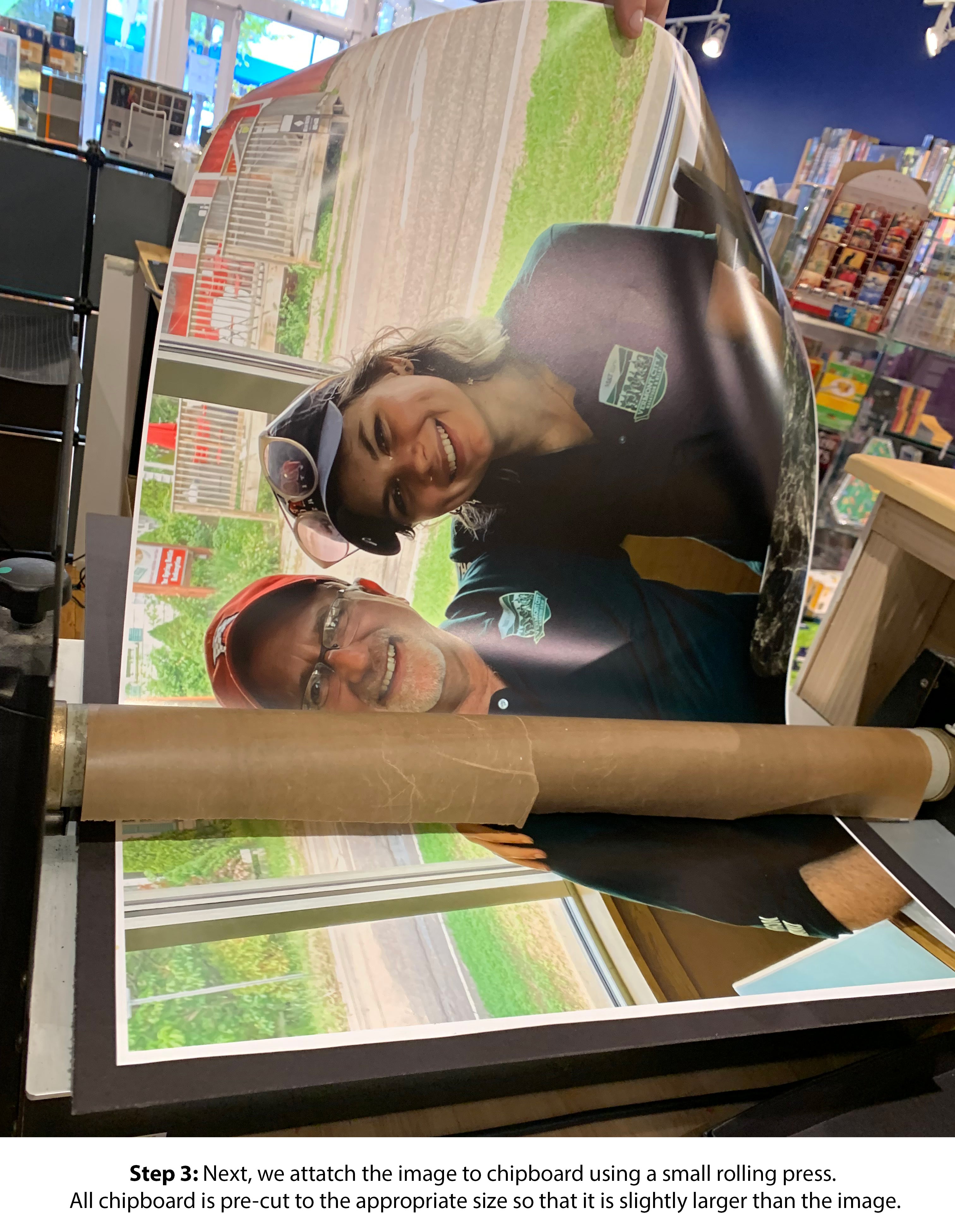 A step in the process of creating a custom jigsaw puzzle. A vibrant image is being attached to chipboard using a small rolling press. The image features two people smiling. The chipboard is pre-cut for a perfect fit, ensuring a quality jigsaw puzzle experience.