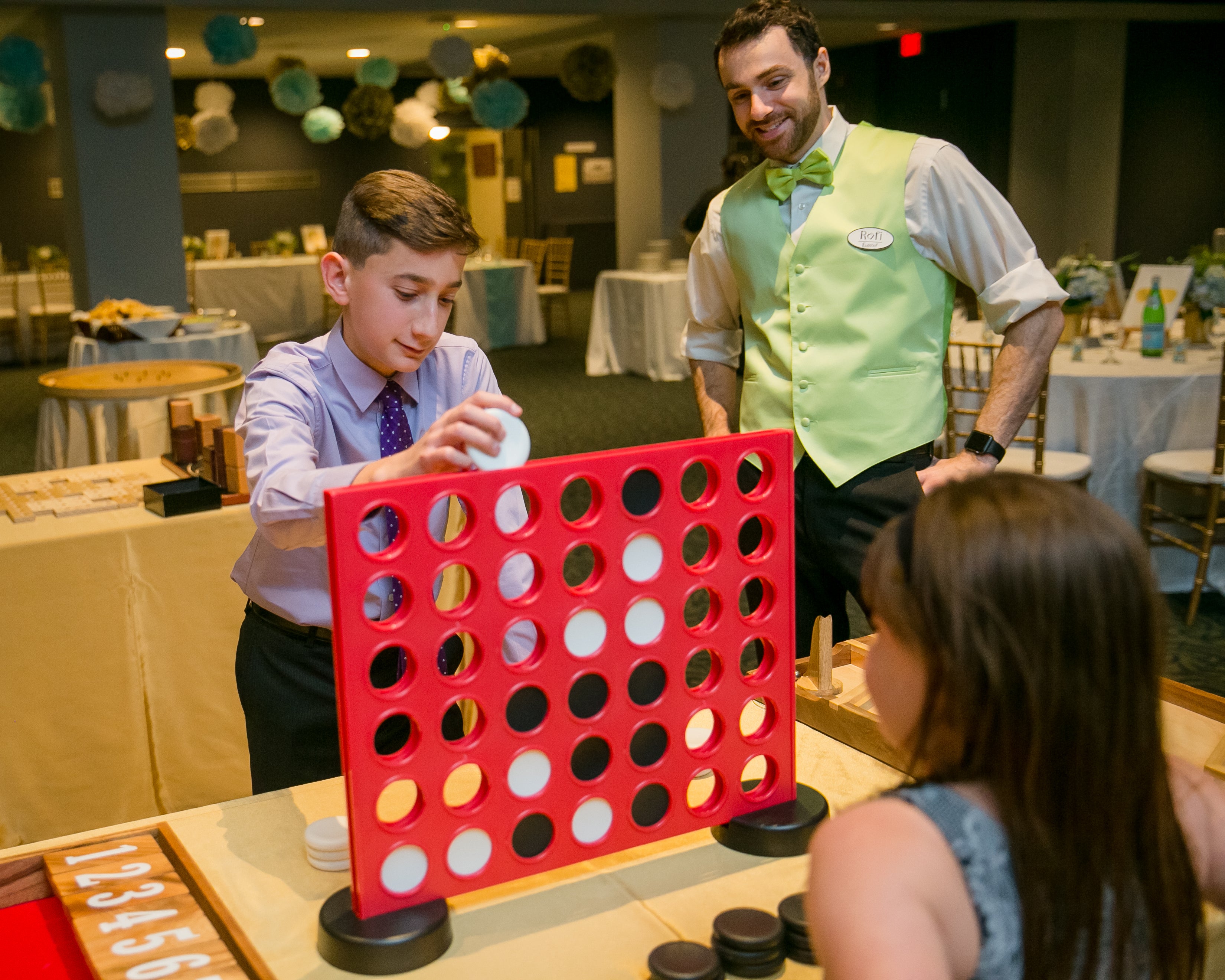 Two children play Connect Four on a large red board.  A man in a green vest watches. 