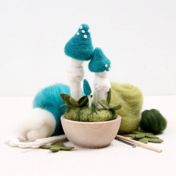 A Blue Roundhead Needle Felting Kit from BEnzie Design featuring a wooden bowl filled with green felted moss and two blue mushrooms with white tops. The kit includes assorted wool in blue, green, and white, along with felting needles and leaf-shaped felt pieces for crafting.