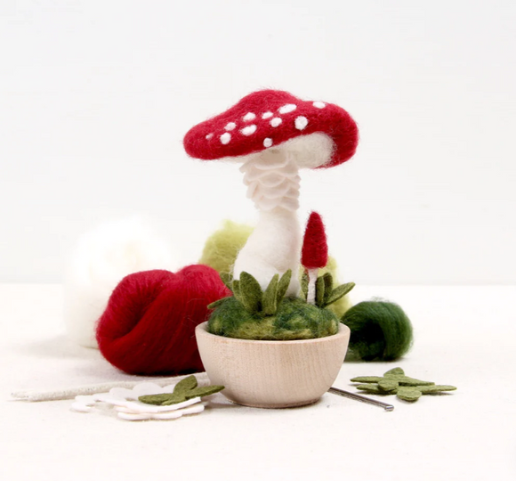 The Crimson Toadstool Needle Felting Kit features colorful wool roving in shades of red, white, and green, and is centered around a charming red and white toadstool mushroom sitting atop a green felt base in a wooden bowl. The kit includes various materials for crafting a delightful mushroom scene.