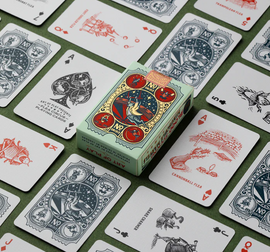 Flea Circus Playing Cards by Art of Play displayed on a green surface. The box features vibrant illustrations of a circus scene with various fleas depicted on the cards, showcasing unique artwork and playful themes.