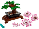 LEGO® Bonsai Tree DIY kit featuring a detailed tree with lush green leaves and pink flowers. The bonsai is displayed in a black pot on a wooden stand, with additional pink flower pieces scattered around for customization. Includes a blue tool for assembly.