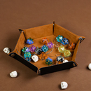 A hex-shaped game tray made of camel hex leatherette and velvet, featuring an assortment of colorful dice including d20s and d12s. The interior is a plush velvet finish in camel color, while the edges are sleek black leatherette. Small decorative items resembling carved stones are scattered around the tray, creating a cozy gaming setup.