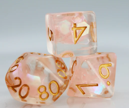 A close-up view of the Nixie Nimbus RPG Dice Set by Foam Brain, showcasing three beautifully crafted dice in a light pink color with shimmering features and golden numbering. Perfect for tabletop RPG gaming, these dice combine style and functionality.