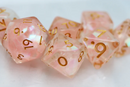 A close-up view of the Nixie Nimbus RPG Dice Set featuring translucent pink dice with gold numbering. The set includes various polyhedral dice, perfect for tabletop RPG games. The shimmering surface adds an enchanting touch to gaming sessions.
