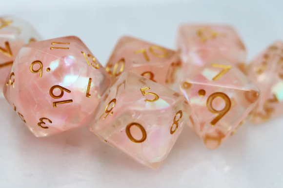 A close-up view of the Nixie Nimbus RPG Dice Set featuring translucent pink dice with gold numbering. The set includes various polyhedral dice, perfect for tabletop RPG games. The shimmering surface adds an enchanting touch to gaming sessions.