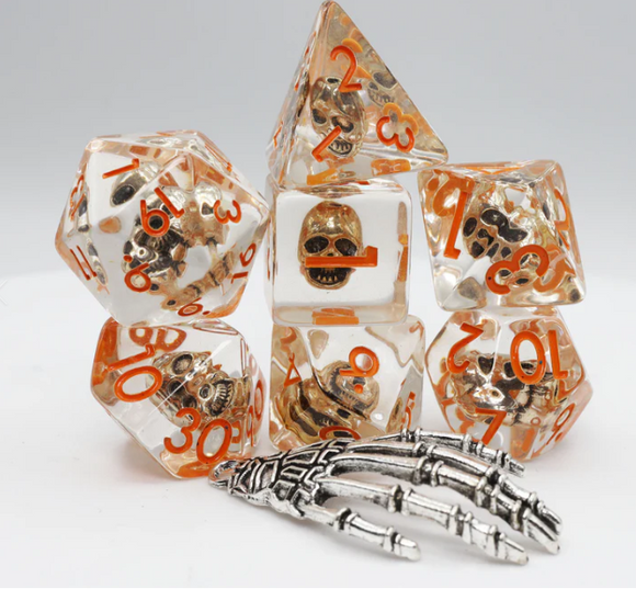 Laughing Skull RPG Dice Set features a collection of seven polyhedral dice. Each die is encased in transparent resin with intricate skull designs and bright orange numbers. A metallic silver skeleton hand is placed next to the dice, adding to the unique theme. Perfect for tabletop role-playing games, these dice are designed for both aesthetics and functionality.