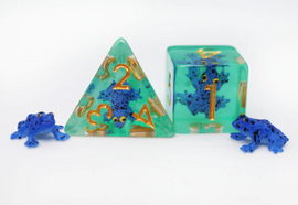 A set of dark blue frog-themed RPG dice made by Foam Brain. The set includes a green d20 die with blue frogs and gold numbers, a green d4 die with similar designs, and two small blue frog figurines beside them. The dice feature a unique water-like appearance with vibrant colors, ideal for tabletop games.
