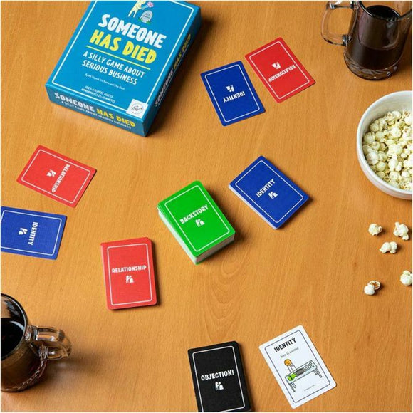 Someone Has Died game box in blue on a wooden table. Surrounding the box are various game cards in red, blue, and green, labeled with terms like 'Identity,' 'Relationship,' and 'Backstory.' A bowl of popcorn and a mug of coffee are also present.
