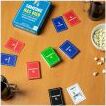 A tabletop game titled 'Someone Has Died' by Gather Round Games. The game is displayed in a blue box, surrounded by colorful cards in red, blue, green, and black laid out on a wooden table. Snacks and a cup are also visible, creating a cozy game night atmosphere.