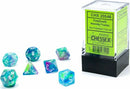 Festive mini polyhedral 7-die set from Chessex in vibrant colors including blue, green, and purple. The dice feature clear numbers on each face and are displayed beside their transparent packaging.