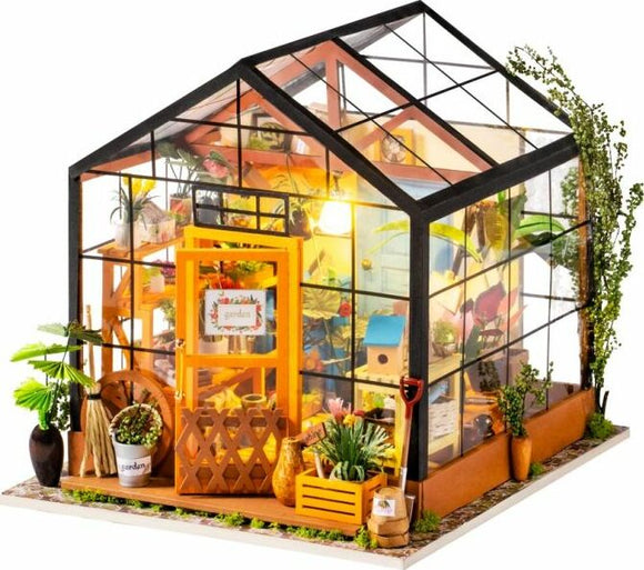 Cathy's Flower House Model Kit displayed prominently. The kit features a detailed miniature greenhouse with vibrant plants, a bright yellow door, and decorative garden items, showcasing a charming garden scene perfect for DIY enthusiasts.