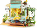 A detailed model kit of a quaint sweet shop called 'Flowery Sweets & Teas'. The model features a vibrant storefront with a sign, decorative flowers, a small outdoor seating area with a yellow checkered tablecloth, and a charming swing. The scene captures a whimsical atmosphere perfect for any DIY enthusiast.