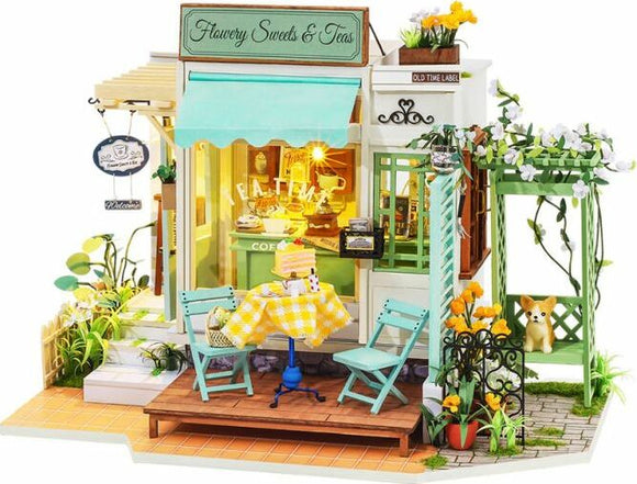 A detailed model kit of a quaint sweet shop called 'Flowery Sweets & Teas'. The model features a vibrant storefront with a sign, decorative flowers, a small outdoor seating area with a yellow checkered tablecloth, and a charming swing. The scene captures a whimsical atmosphere perfect for any DIY enthusiast.