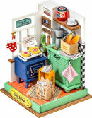 A colorful DIY kit from Robotime titled 'DIY Afternoon Baking Time.' The scene features a quaint kitchen with a blue stove displaying heart-shaped cookies, a green cupboard filled with boxes of ingredients, and a basket of fresh bread. Decorative elements include a window with a sign that reads 'Bread and Cappuccino,' hanging mitts, and various kitchen utensils, creating a charming baking atmosphere.