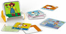 The Dress Code game by Smart Toys & Games features a fun, interactive mechanical puzzle set. It includes a stand with a colorful cartoon character holding a flower, accompanied by various illustrated cards showing different outfits and activities. The cards are placed around the stand, displaying playful images of children in seasonal clothing, engaging in sports, and enjoying various activities.